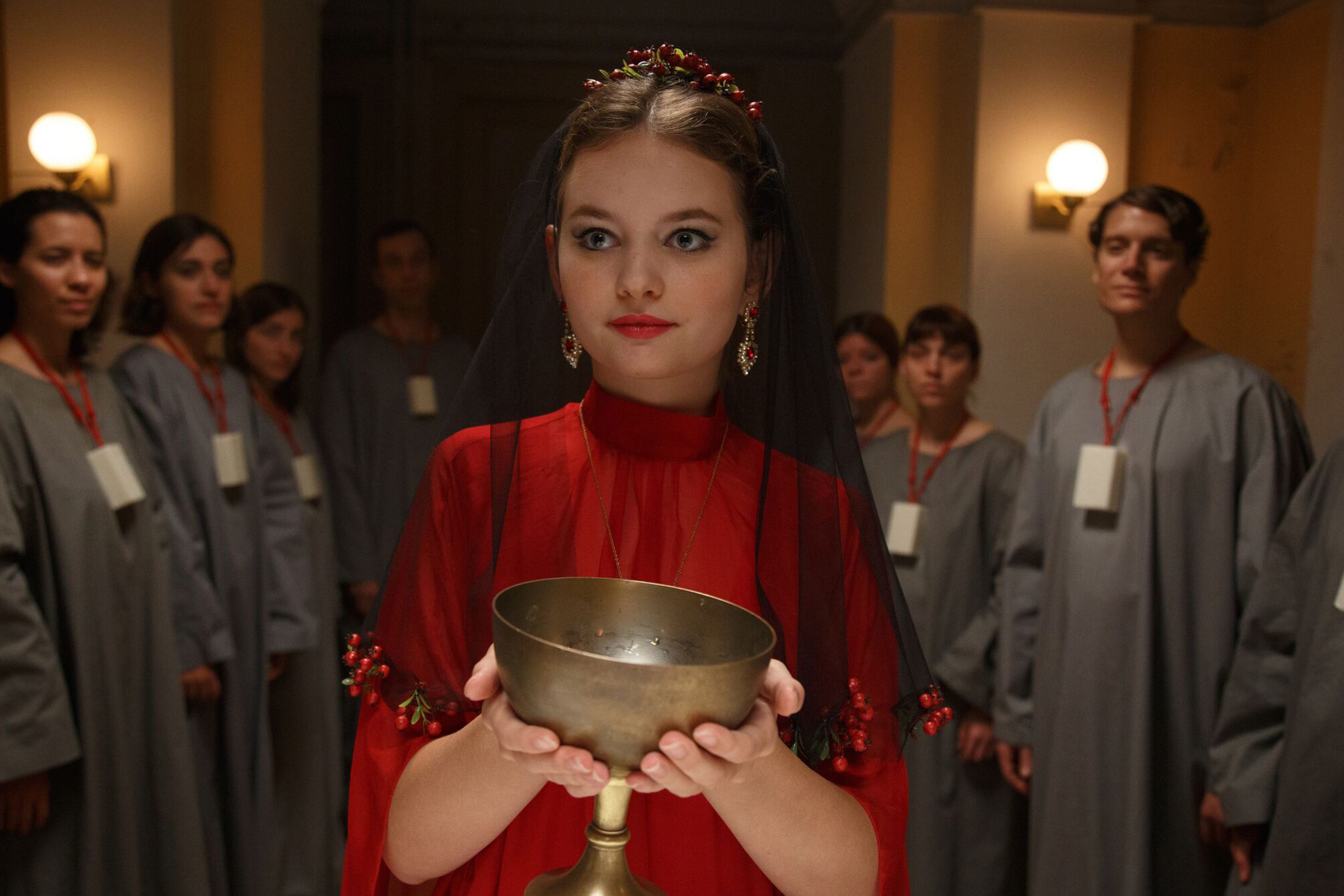 Irina (Greta Bohacek) in einem roten Kleid und einer Schale aus Bronze in ihren Händen.